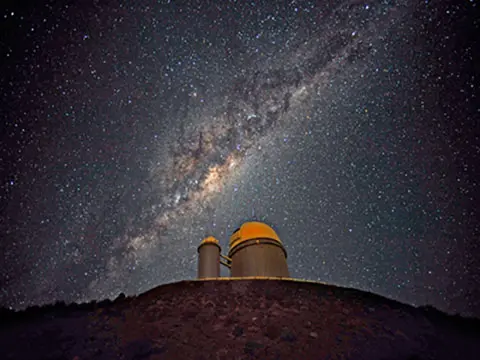 The ESO 3.6-metre telescope at La Silla, during observations. Across the plan of the picture, is the Milky Way, our own galaxy, a disc-shaped structure seen perfectly edge-on. Above the telescope´s dome, here lighted by the Moon, and partially hidden behind dark dust clouds, is the yellowish and prominent central bulge of the Milky Way. - Original Infinities by Lauren Camp